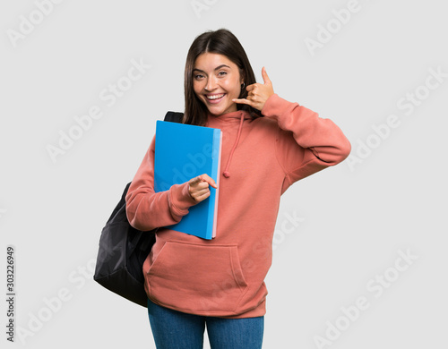 Young student woman holding notebooks making phone gesture and pointing front over isolated grey background