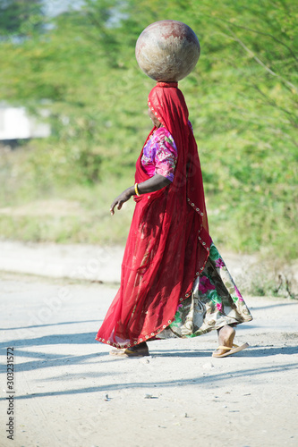 traditionally dressed woman in traditional indian Saree walking at rural road with pot at head. Rajasthan. India