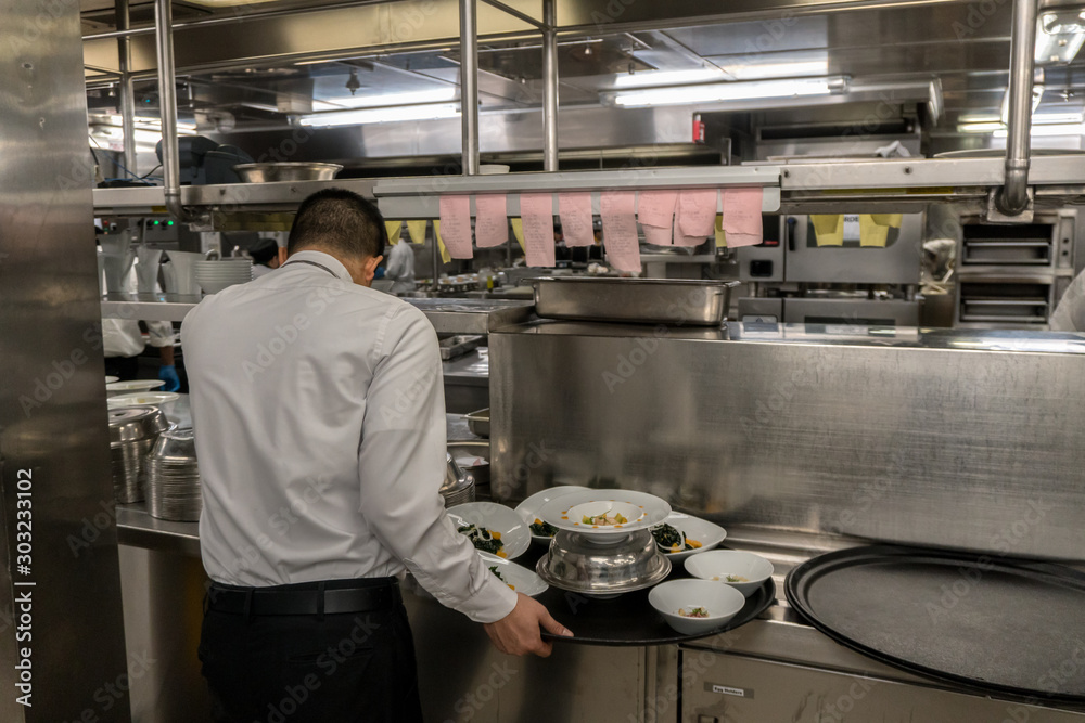 Waiter in the uniform is waiting for his orders. Big galley on a cruise ship. Black tray and a lot of plates around. Waiter in the uniform is waiting for his orders. Big galley on a cruise ship. Black tray and a lot of plates around.