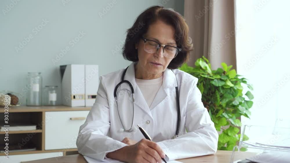 60s American female doctor talking and writing, working at table in clinic office interior. 4k Portrait of serious mature expert speaking and doing paperwork sitting in interior. Concept: healthcare 60s American female doctor talking and writing, working at table in clinic office interior. 4k Portrait of serious mature expert speaking and doing paperwork sitting in interior. Concept: healthcare