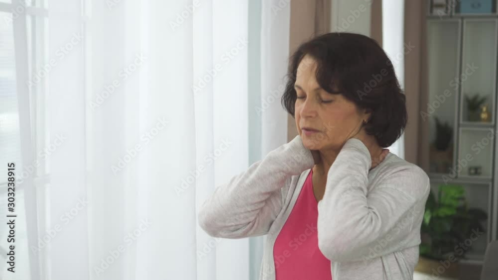Elderly american woman massage neck with hand standing in apartment room. Tired mature female touching shoulder to relieve back pain and stress. Retired 60s lady having health time in house interior Elderly american woman massage neck with hand standing in apartment room. Tired mature female touching shoulder to relieve back pain and stress. Retired 60s lady having health time in house interior