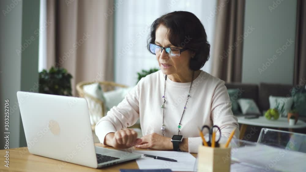 Mature woman accountant working with laptop, writing at table in home office. American aged businesswoman watching computer screen and doing work with financial document at desk in room. Beautiful 60s Mature woman accountant working with laptop, writing at table in home office. American aged businesswoman watching computer screen and doing work with financial document at desk in room. Beautiful 60s