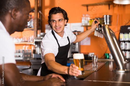 Positive man bartender giving glass of golden beer to man client