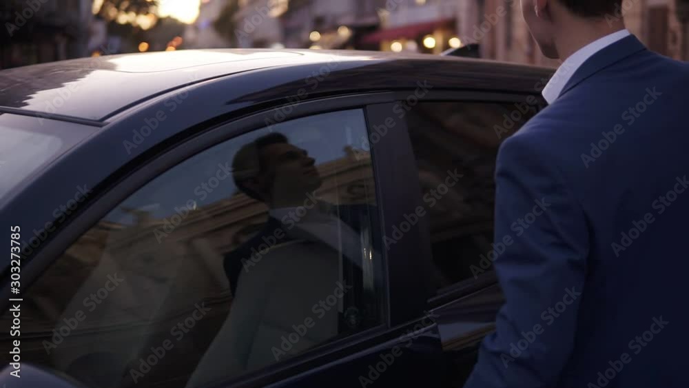 Slow motion young man walk sitting in the black car on street. Smiling businessman in elegant blue suit opens the door of his expensive car and sitting into. Excited close the door. Evening city dusk