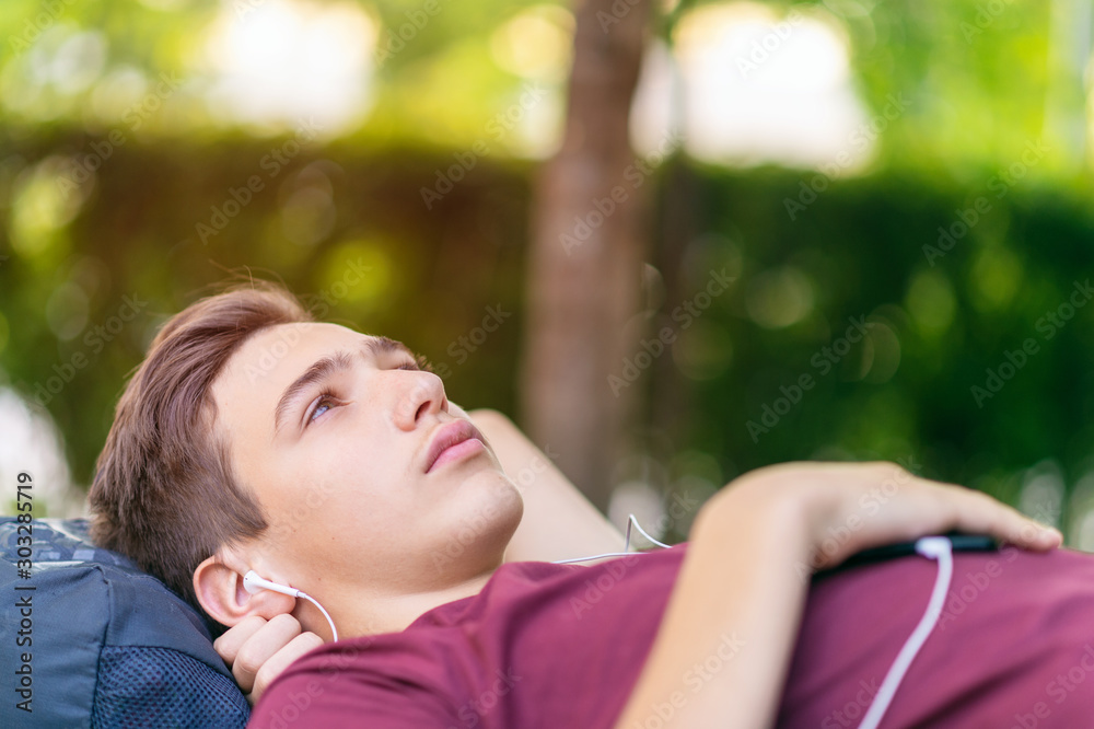 Close-up portrait of a sad young man lying on the bench in the park.  Upset teenage boy relaxing outdoors. Teenager is lying on the nature with a loneliness feelings. Soft focus effect.