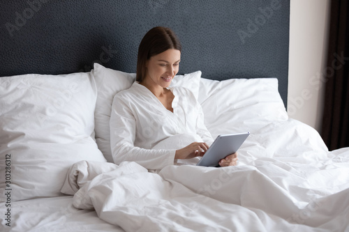 Happy young woman using digital tablet computer sitting in bed