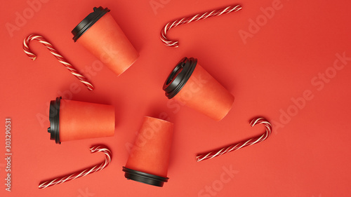 Paper coffee cup with sugar canes on a red background. Christmas background with Xmas candy canes and cofee. Top view, flat lay, mockup. Copy space for text.