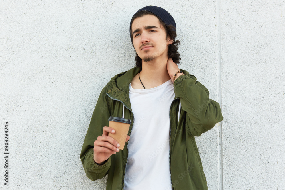 Fototapeta premium Photo of upset disappointed youngster looking aside, touching his neck with hand, feeling pain, being very tired, having lunchtime, spending leisure time alone, wearing casual items of clothes.