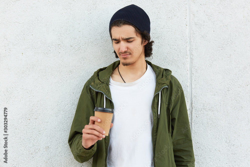 Fototapeta premium Portrait of confused unsatisfied hipster standing isolated over white wall outdoors, holding papercup with drink, undelighted with taste of drink, wearing casual clothes. Street style concept.