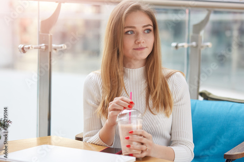 Image of dreaming tender lady looking aside, having pleasant facial expression, holding cup with coctail, sitting alone in cafe, enjoying free time, wearing sweater. People and free time concept. photo