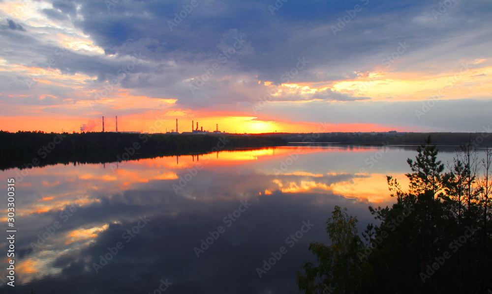 Heavy clouds at sunset hang over a quiet river. Reflection of the sunset and clouds in the quiet surface of the river. 