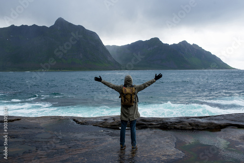 A man with backpack walking along an empty ocean beach and looking at the mountains. Fjord in Norway. Scenic view. Travel, adventure. Sense of freedom, lifestyle. Lofoten Islands, Norway. North summer
