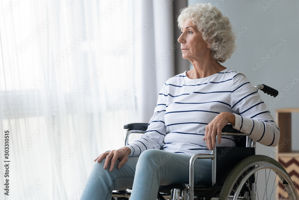 Sad paraplegic old woman sit on wheelchair look through window - obrazy, fototapety, plakaty 
