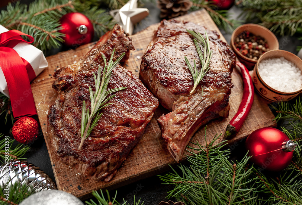 Christmas dinner for two, grilled beef steak ribeye, herbs and spices on a stone table with a Christmas tree and New Year's toys - obrazy, fototapety, plakaty 