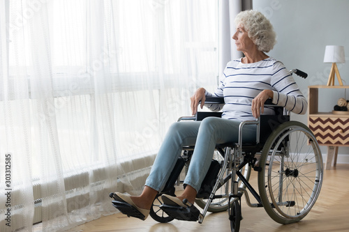 Pensive disabled senior grandma sit on wheelchair look through window