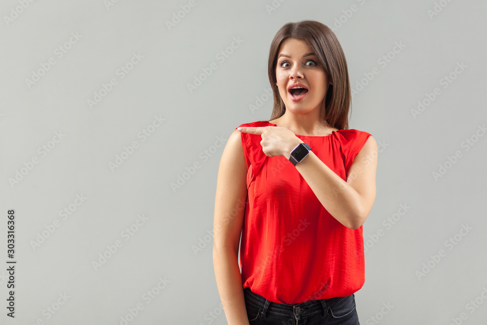 Portrait of shocked beautiful brunette young woman in red shirt standing, looking, amazed surprised face and pointing at empty background copyspace. indoor, studio shot, isolated on gray background. Portrait of shocked beautiful brunette young woman in red shirt standing, looking, amazed surprised face and pointing at empty background copyspace. indoor, studio shot, isolated on gray background.