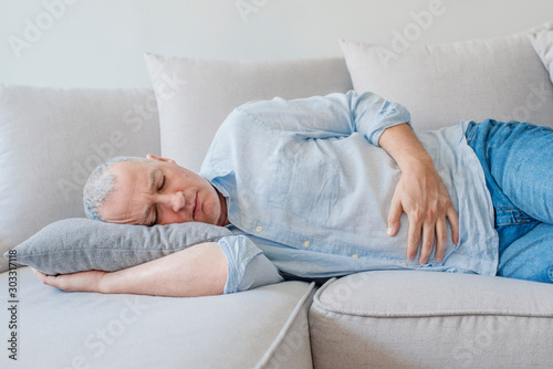 Terrible stomachache. Frustrated handsome young man hugging his belly and keeping eyes closed while lying on the couch at home