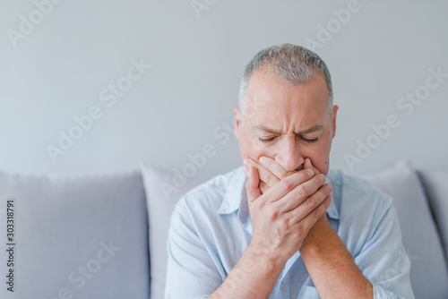 Toothache. Frustrated young man touching his cheek and keeping eyes closed while sitting on the couch at home