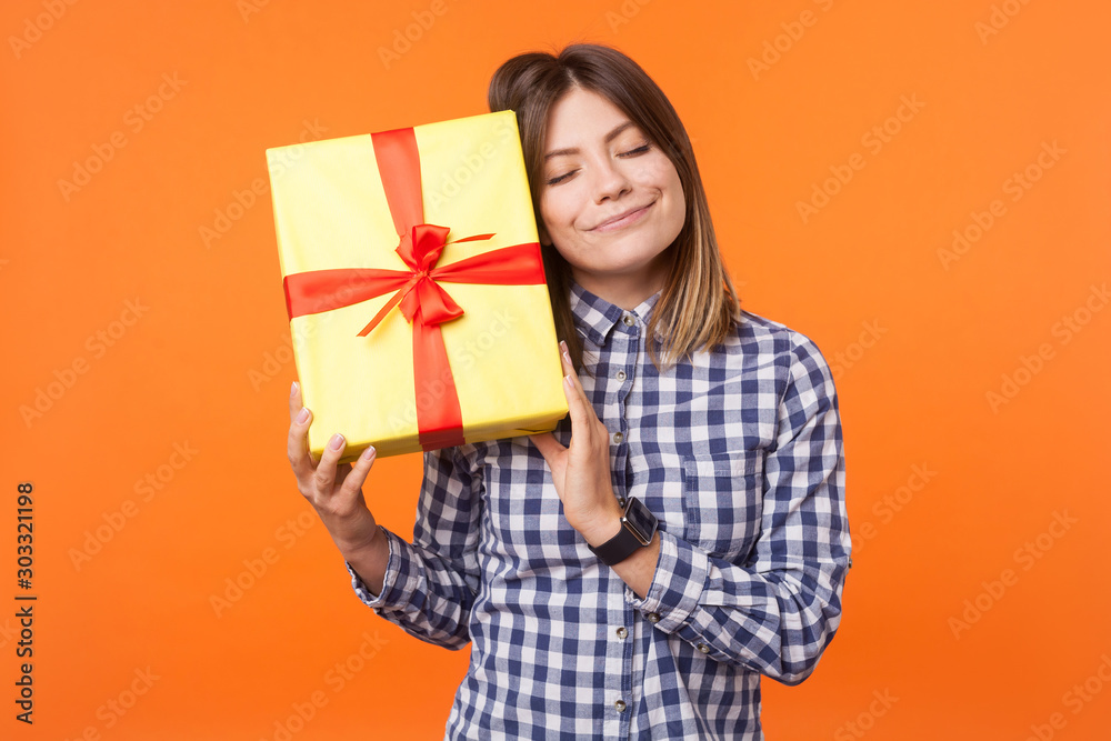 Portrait of dreamy woman with charming smile in casual checkered shirt standing with closed eyes, holding gift box near head, full of dreams and hopes. indoor studio shot isolated on orange background Portrait of dreamy woman with charming smile in casual checkered shirt standing with closed eyes, holding gift box near head, full of dreams and hopes. indoor studio shot isolated on orange background