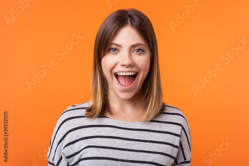 Portrait of surprised happy woman with brown hair in long sleeve shirt standing, looking with amazement and opened mouth, unexpected positive news. indoor studio shot isolated on orange background