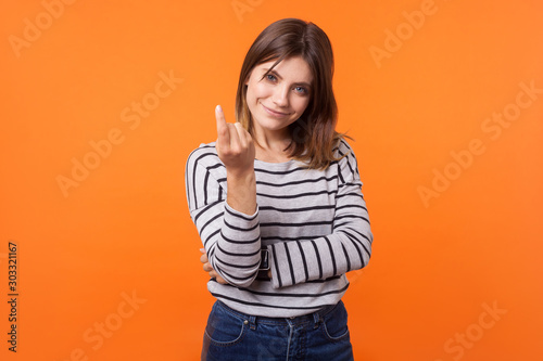 Photos Portrait of friendly pretty young woman with brown hair in long sleeve striped shirt standing, beckoning with finger to come nearer, smiling at camera