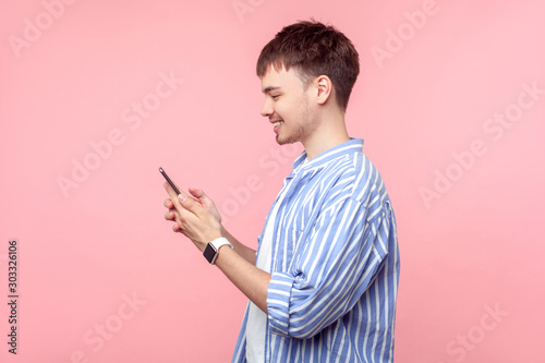 Side view of happy young brown-haired man with small beard in casual striped shirt holding phone, smiling while browsing social media, reading message. indoor studio shot isolated on pink background
