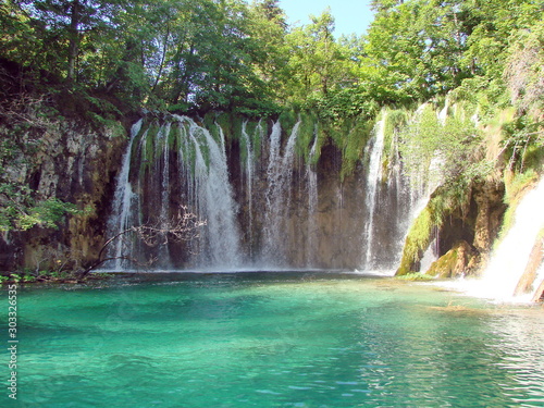 Panorama of waterfalls flowing into the lake surrounded by mighty mountain forest trees on a clear sunny day.