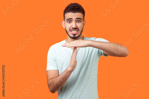 I need more time! Portrait of frustrated brunette man with beard in casual white t-shirt showing time out gesture, looking with imploring eyes, hurry to meet deadline. studio shot on orange background