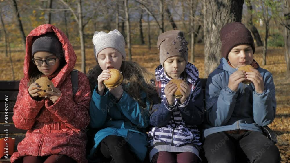 Group of hungry kids are eating burger outdoor in the park in sunny autumn day Group of hungry kids are eating burger outdoor in the park in sunny autumn day