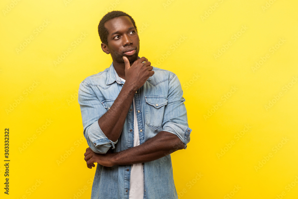 Thinking over decision. Portrait of pensive smart handsome man in denim casual shirt looking up, holding his chin while considering problem solution. indoor studio shot isolated on yellow background - obrazy, fototapety, plakaty 