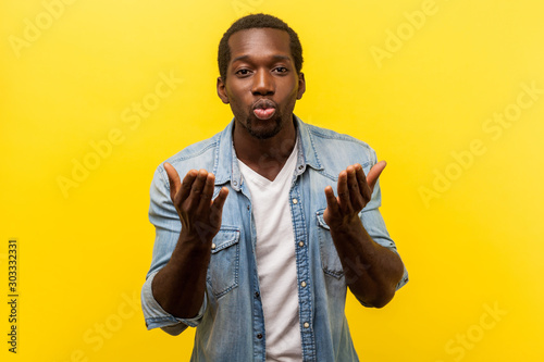 Portrait of happy handsome man in love wearing denim casual shirt with rolled up sleeves, sending air kiss at camera, sharing romantic feelings. indoor studio shot isolated on yellow background
