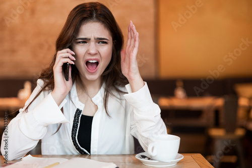 young woman in airphones having unpleasant phone conversation working outside office in her laptop during lunch break drinking hot coffee in cafe looking irritated photo