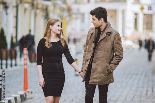 Urban modern young love couple walking romantic talking talking  holding hands on a date. Young multicultural Turkish brunette and Caucasian couple on old european street. Autumn spring weather