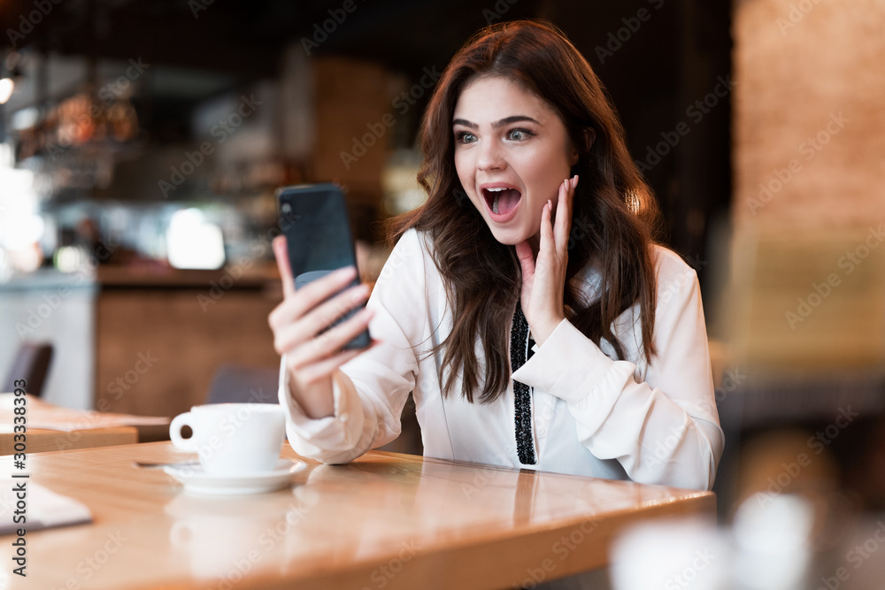 young beautiful woman in white stylish jacket with smartphone in her hands looking surprised while speaking with friend during coffee break in cafe modern businesswoman young beautiful woman in white stylish jacket with smartphone in her hands looking surprised while speaking with friend during coffee break in cafe modern businesswoman