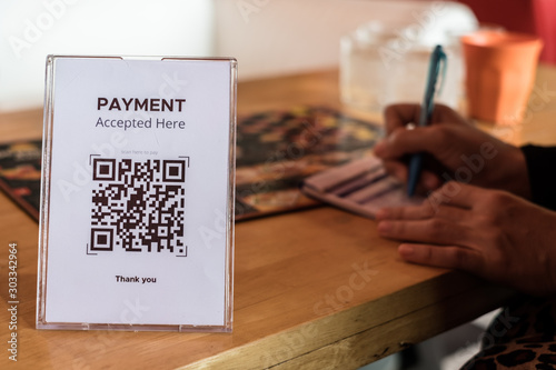 QR code payment accepted in acrylic or plexiglass table card holder on wooden table for restaurant with hand writing down food menu. Mobile banking, commerce, finance technology.