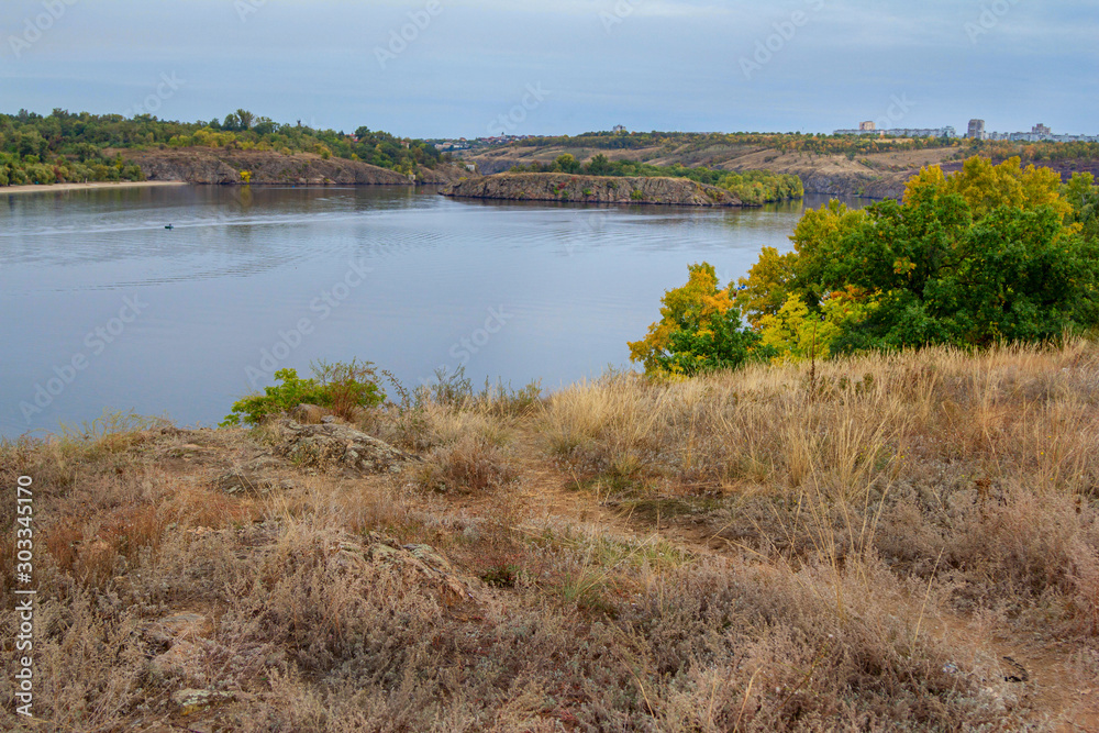 Autumn landscape with calm river, dry grass, cliffs and colorful – green, orange and yellow foliage on the trees. Fall nature and blue sky.