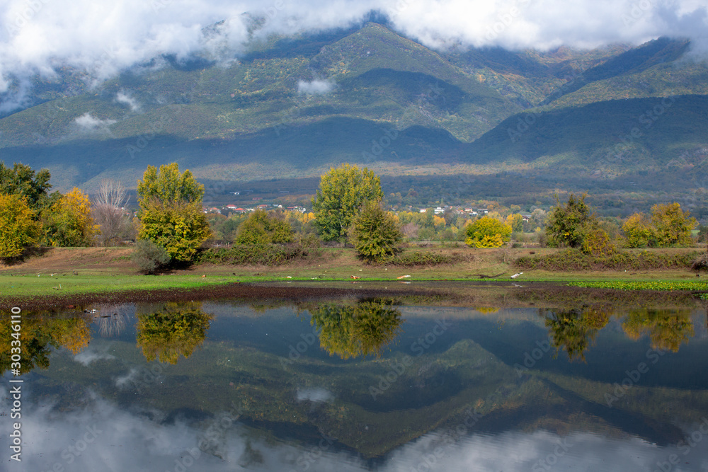 Fototapeta premium Autumn landscape. Trees, mountain hills and cloudy sky reflected in the river. Megalochori, Serres, Greece.