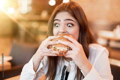 young woman in white stylish blouse biting with appetite fresh tasty meat burger during lunch in trendy cafe looking very hungry eating outside