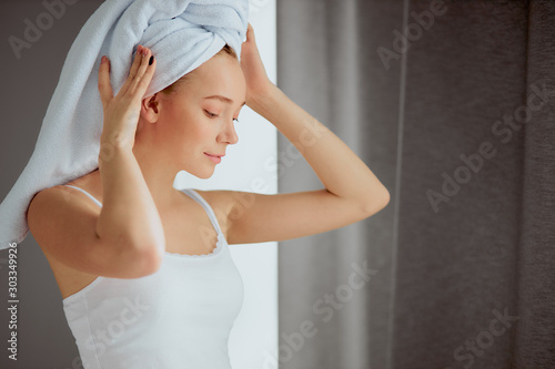 Cute attractive female after bath, standing in front of window with white hotel towel on head, looking down, keeping hands on head, resting after shower, beauty and health treatment concept