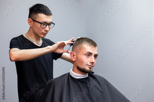 handsome guy has a haircut at the hairdresser, a young Kazakh hairdresser cuts manually with scissors and a comb, the master makes a short haircut against a gray wall