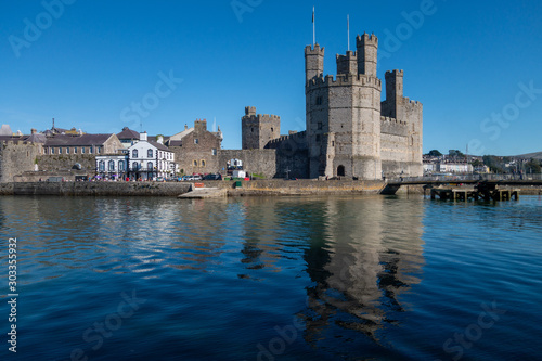 Caernarfon Castle guards the old walled town of Caernarfon and overlooks the river Seiont flowing into the Menai Strait on a clear and sunny day. The Castle is reflected in the high tide.