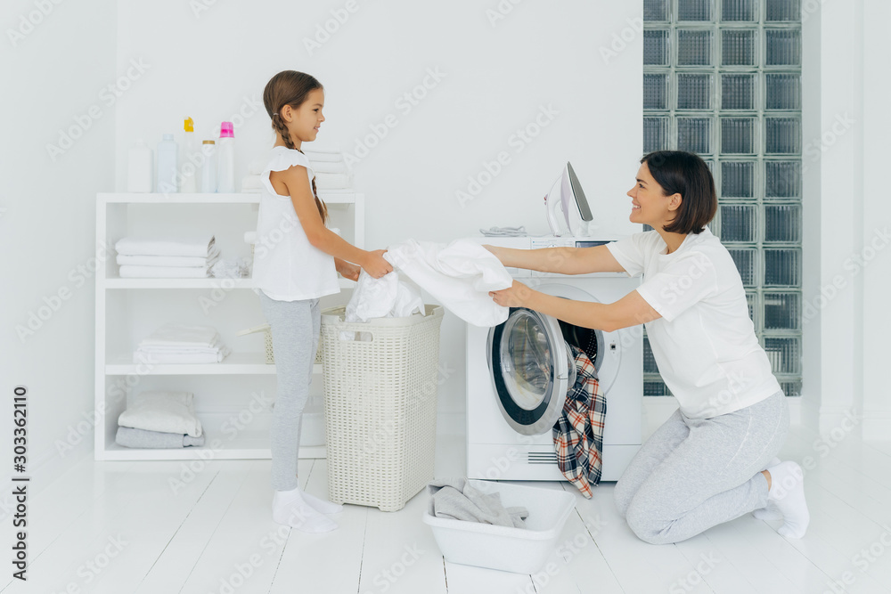 Φωτογραφία Happy mother loads clothes in washing machine, little girl helps, gives white linen from basket, do housework together, dressed in casual clothing