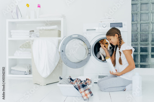 Small child plays with dog russell terrier  poses on knees near washing machine  busy with housekeeping and doing laundry  holds white bottle with washing powder  wears domestic comfortable clothes.