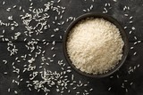 White uncooked, raw long grain rice in black bowl on rustic, black wooden table with copy space top view flat lay from above