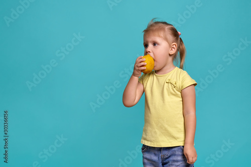 Close-up studio shot of a beautiful little girl. Little blonde girl in a yellow t-shirt on a blue background. The emotions of a child who eats a lemon © samoilova