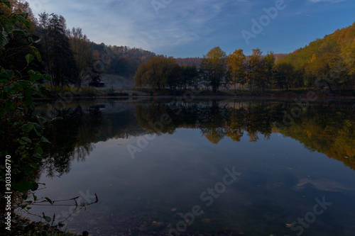 Fresh water lake in a former quarry with reflections of the Indian summer and autumn colors of the leaves on an early morning in November