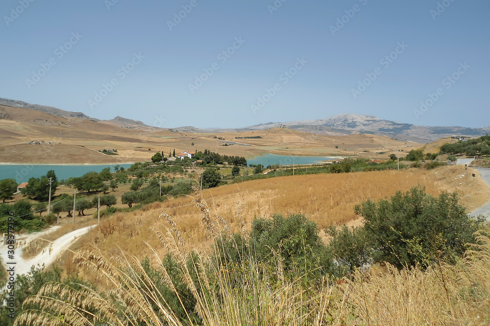 Sicilian landscape with a small villa by a mountain lake in the broiling sun, field with dried grass, green trees, hot summer