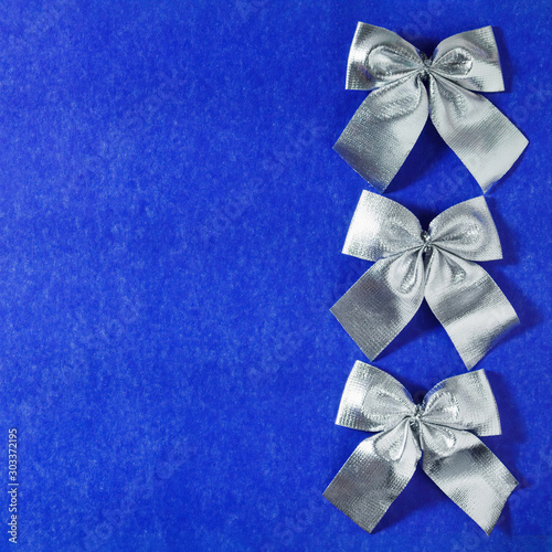 Three silver decorative bows in row on a blue cardboard background. Top view, copy space for text.