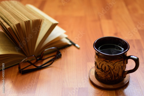 A clay cup with a hot drink on the background of an open book with glasses lying on a sun-drenched wooden table.  Reading a book at breakfast