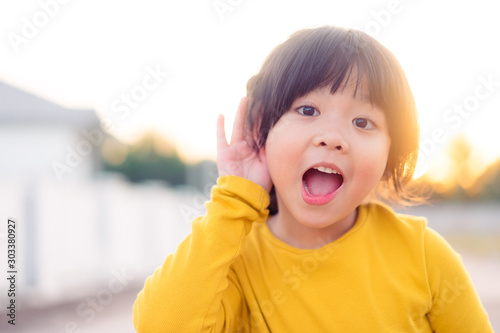 Little asian girl holds her hand near her ear and listening something.Exciting face on funny child girl wear yellow shirt in winter time and listening to curious good news.